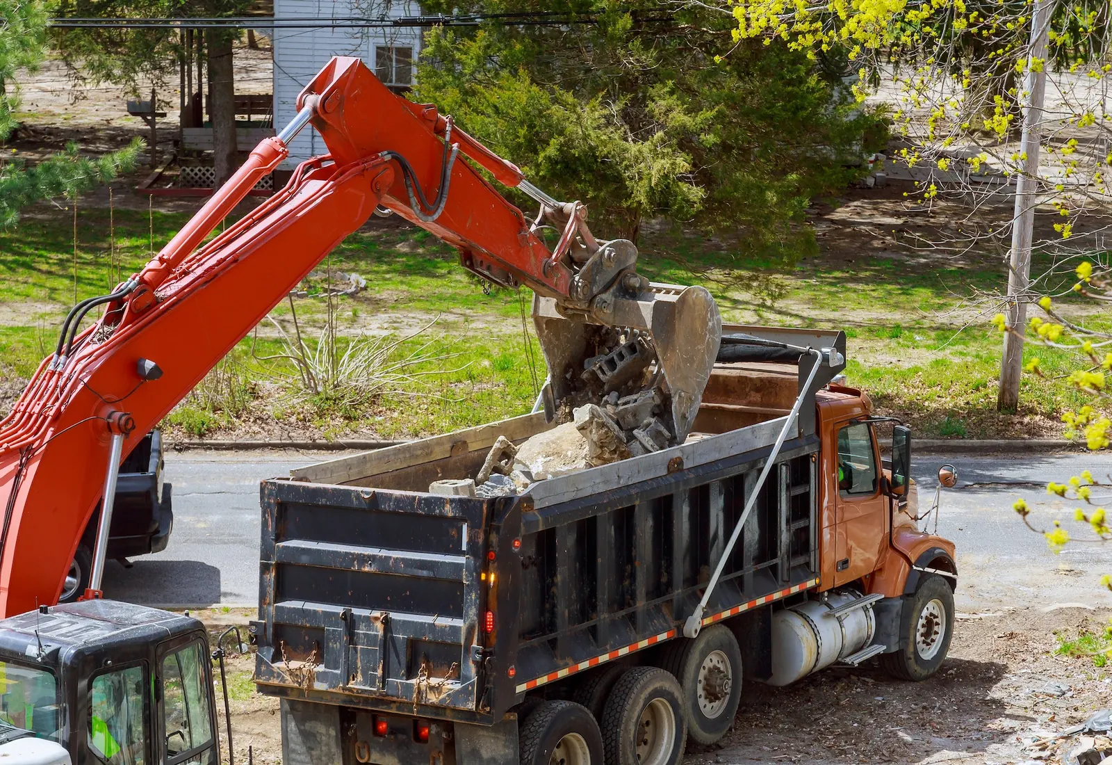 Excavator loading chunks of broken concrete into a dump truck on a residential street