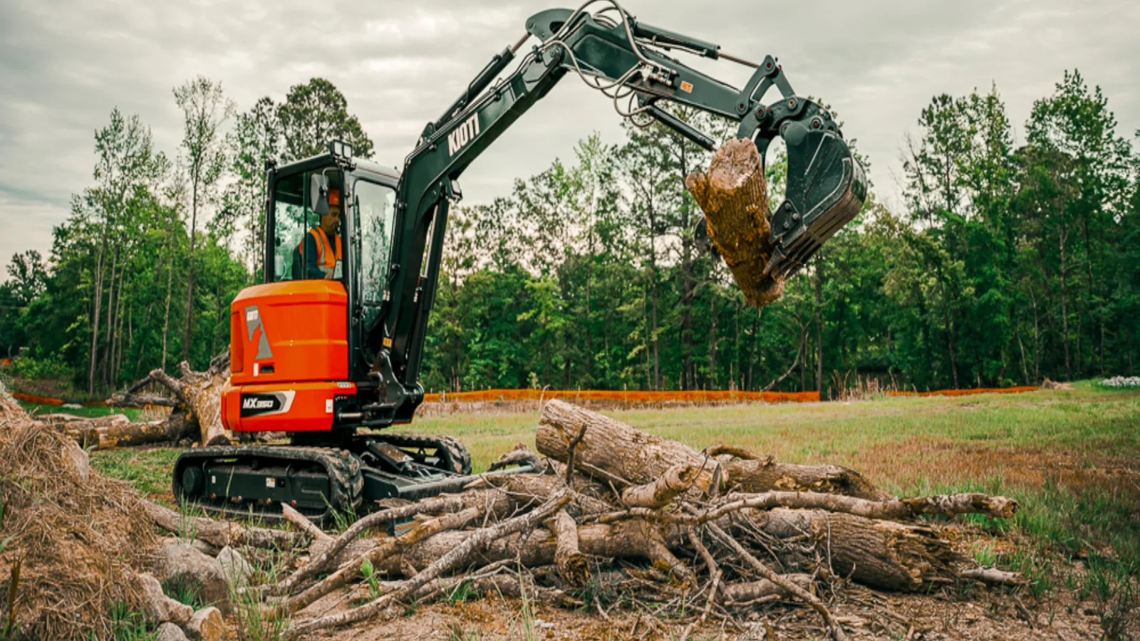 Compact excavator lifting a root wad above cut logs at the edge of a field