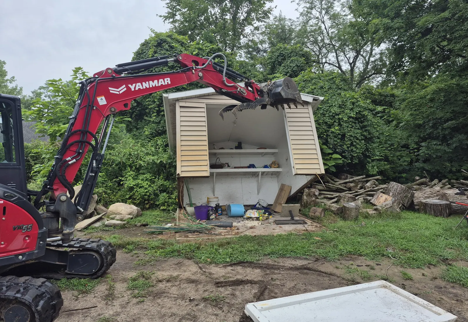 Compact excavator pulling apart a backyard shed beside a woodpile and stone boulders