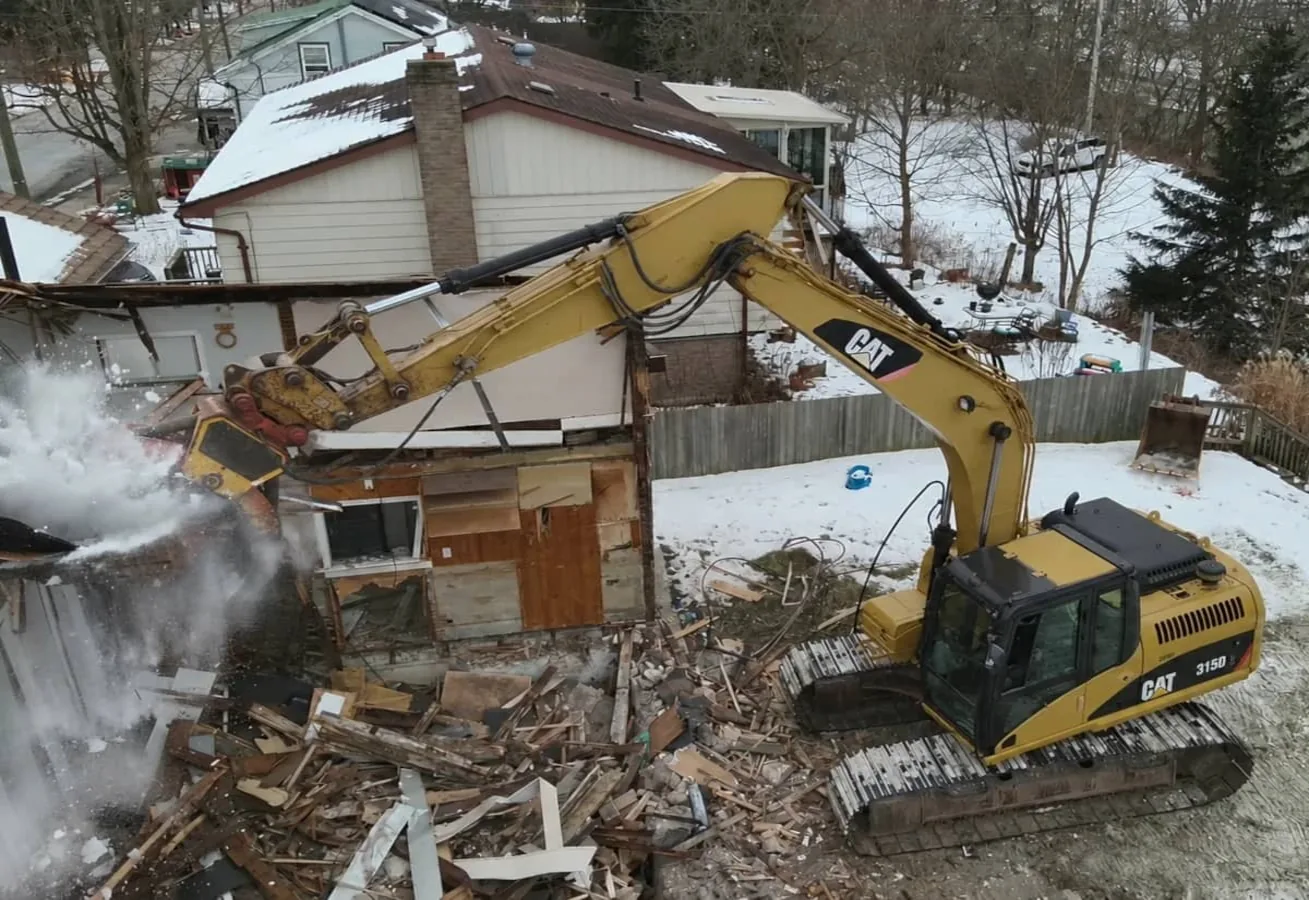 Excavator pulling apart a single-story house in winter, snow on the ground and split lumber below the boom
