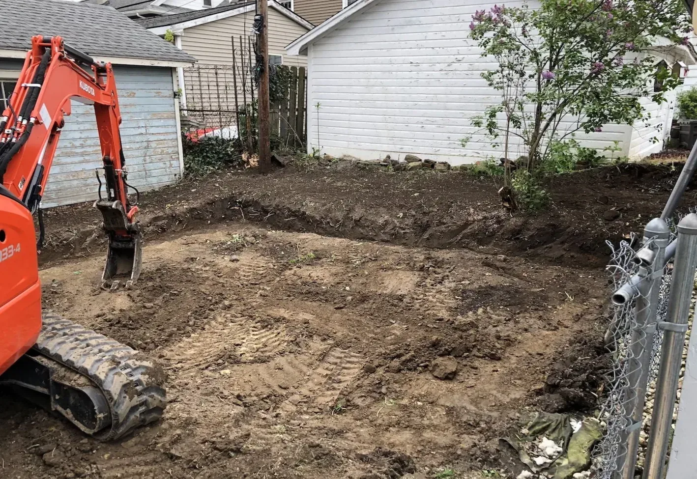 Compact excavator grading a bare dirt yard between a house and a garage