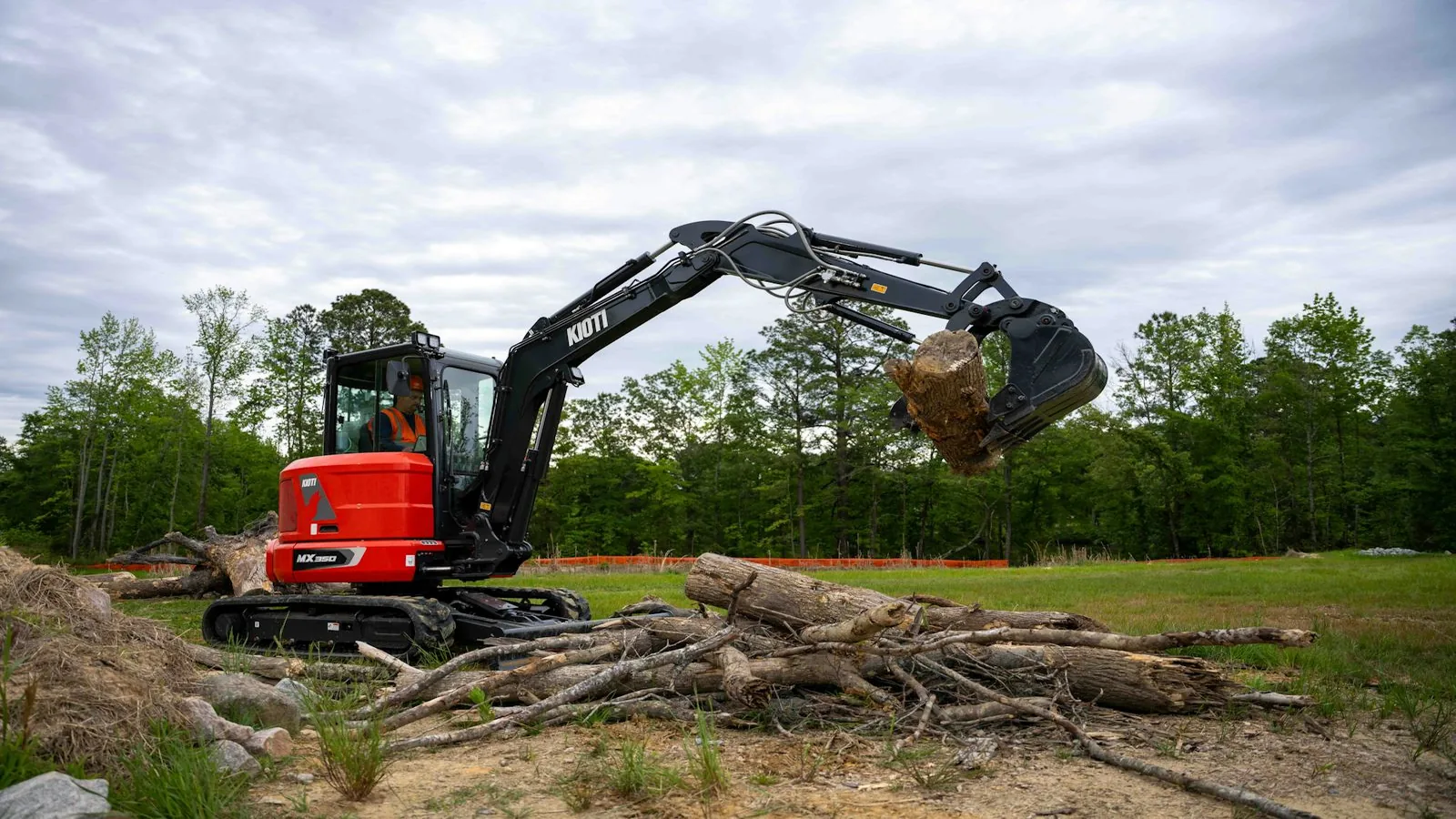 Compact excavator with a grapple lifting a root wad over a pile of logs at a wood line